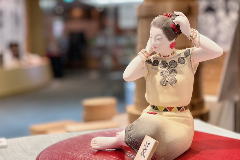 緒方恵子の博多人形の作品「装」です。座っている女性が髪に赤い櫛をつけています。